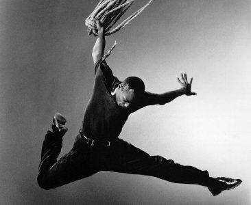 Person in mid-air, holding a rope, wearing dark clothing, against a plain background.
