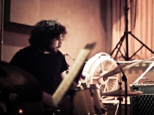 A person with curly dark hair plays drums in a dimly lit room. A fan and music stand are nearby.