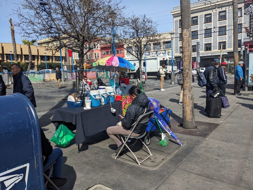 Street vendor with colorful umbrellas and containers at a table in a busy urban setting. People walk by and buildings line the background.