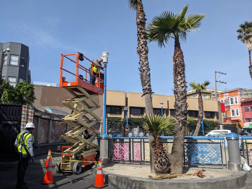 Worker on a scissor lift installing a camera next to a palm tree in an urban area, with construction cones nearby.