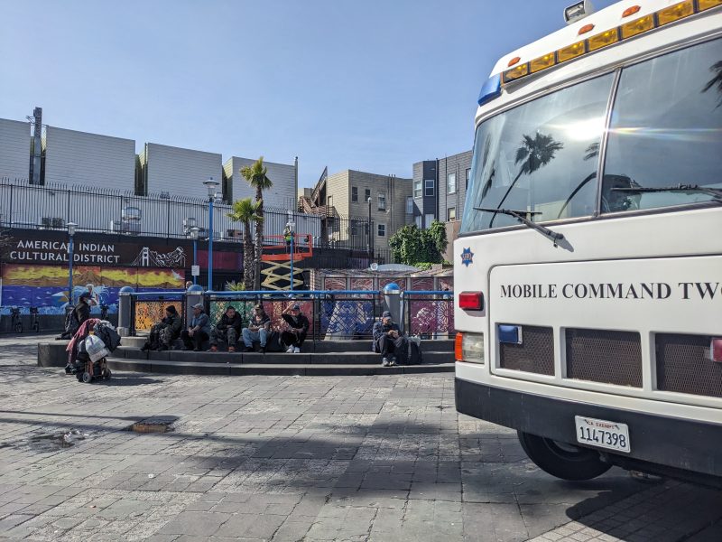 A mobile command truck is parked in a plaza where several people are sitting near a mural. Buildings are visible in the background.