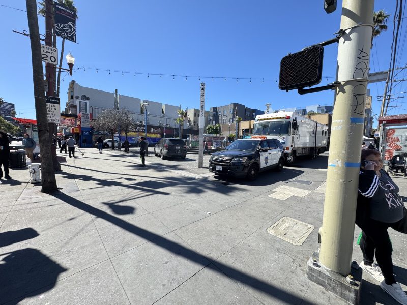 A wide street view shows a police car and a utility truck parked on a city street corner. People walk on the sidewalk under a clear blue sky with string lights above.
