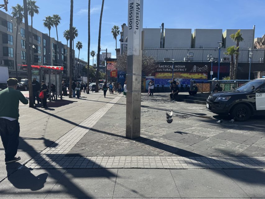 Urban street scene with people walking near a subway entrance. A sign for Mission is visible. A police car is parked nearby, and there is a mural with "American Indian Cultural District" text.