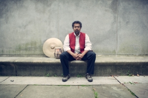 Man sitting on a concrete bench next to a drum, wearing a red vest and white shirt, with a neutral expression against a grey wall background.