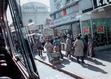 A busy urban street scene with pedestrians walking by shops, including one with a sale sign. A bus window is visible on the left. A building with Leed's Shoes is in the background.