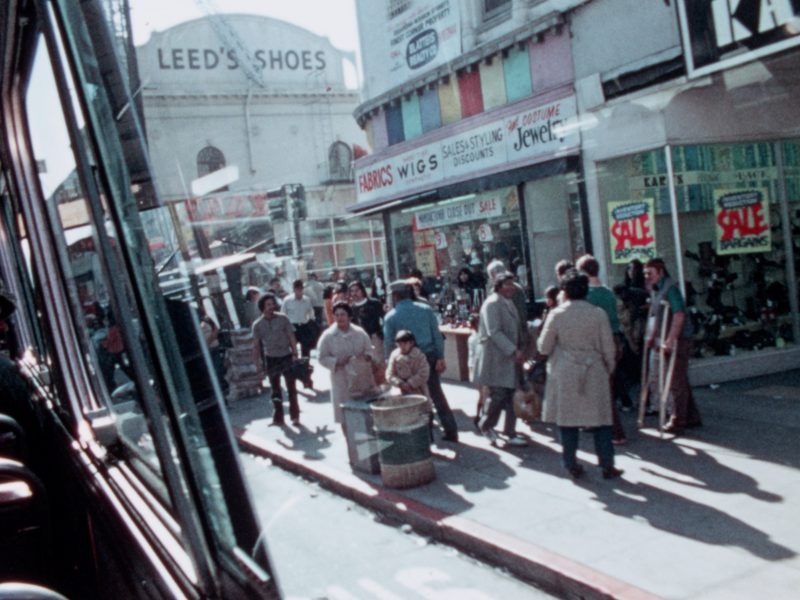 A busy urban street scene with pedestrians walking by shops, including one with a "sale" sign. A bus window is visible on the left. A building with "Leed's Shoes" is in the background.