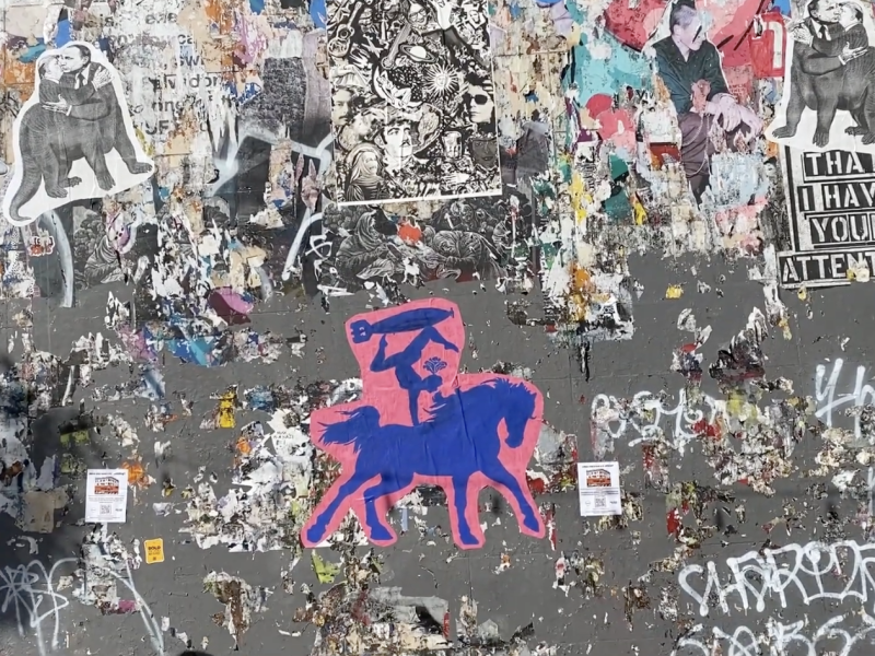 A wall covered with various posters, graffiti, and stickers, including a prominent pink and blue artwork of a rider on a horse. A person is walking past the wall.