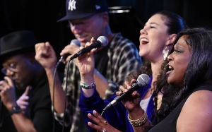 Four people on stage singing into microphones, exhibiting expressions of enthusiasm and engagement.