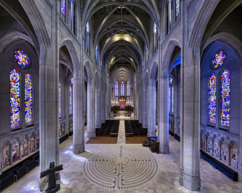 Interior of a grand cathedral with vaulted ceilings, featuring stained glass windows, stone columns, a labyrinth pattern on the floor, and rows of empty pews.