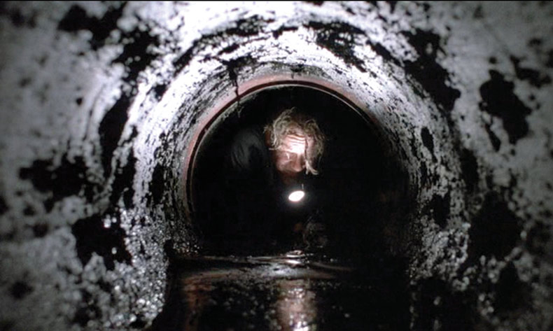 Person with a flashlight crawls through a dark, dirty tunnel.