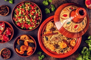 An overhead view of a couscous dish in a decorative pot, surrounded by bowls of salad, spices, dried fruits, and pomegranate seeds on a dark surface.