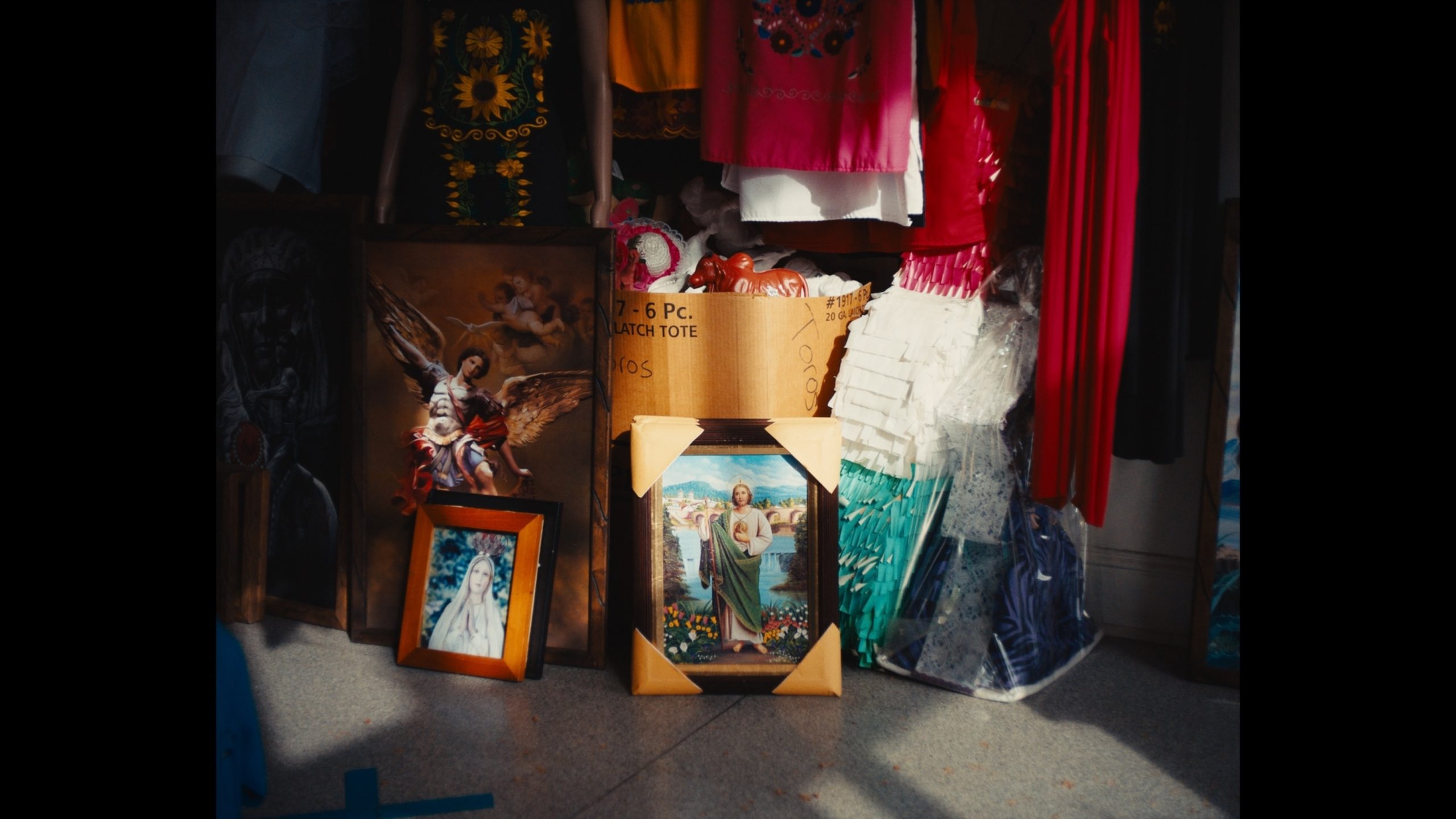 Assortment of religious and decorative items, including framed pictures, colorful clothing, and a cardboard box, arranged on a tiled floor with sunlight streaming in.