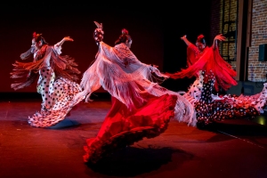 Four dancers performing Flamenco on stage, wearing traditional red and polka dot dresses with flowing fringed shawls, captured mid-movement under stage lighting.