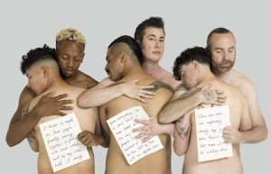 Five people stand close together, embracing. Each holds papers with handwritten text. Their expressions are calm, and they are posed against a plain background.