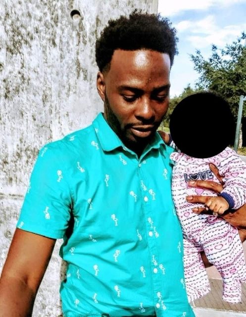 A man in a turquoise shirt stands outdoors, holding a baby dressed in patterned clothing.