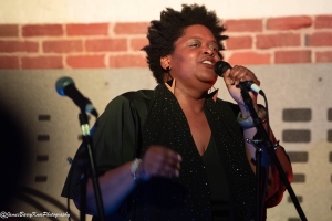 A woman with short, curly hair sings into a microphone, standing in front of a brick wall background.