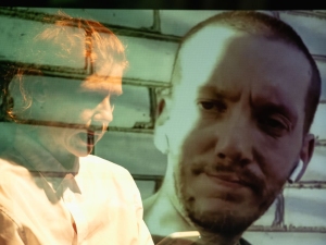 Overlay of two people, one with a serious expression and the other appearing blurred, set against a brick wall background.