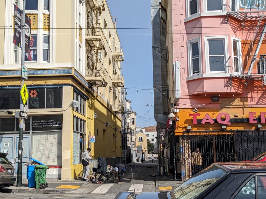 Street view with parked cars, people gathered on a sidewalk, and colorful buildings. A yellow and a pink building flank the alley entrance, and a sign reads "TAQ.