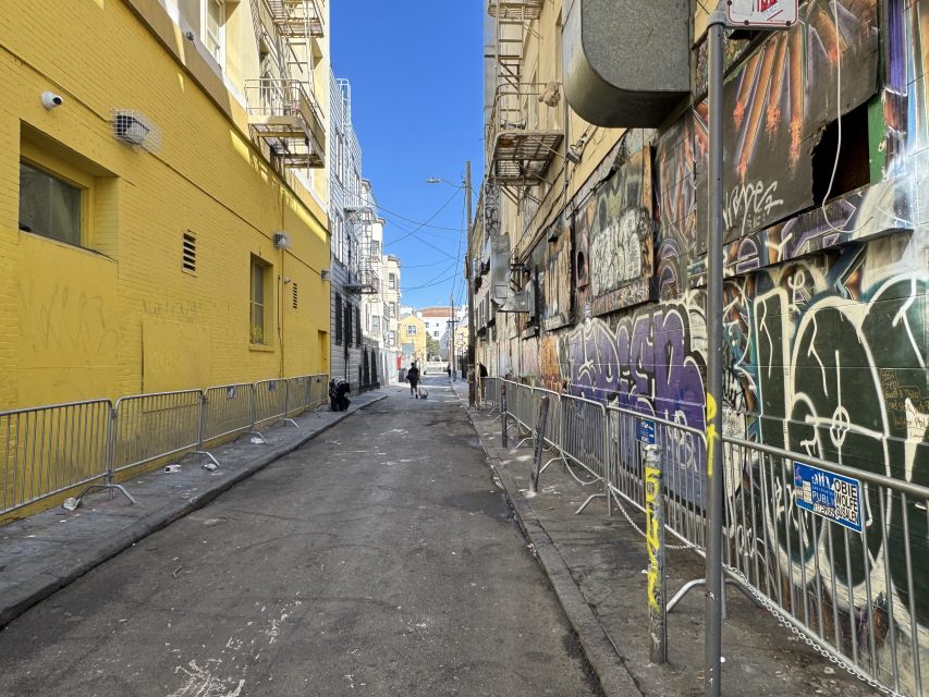 Narrow urban alley with graffiti-covered walls on the right and a yellow building on the left. Metal barricades line both sides. Two people walk in the distance under a clear blue sky.