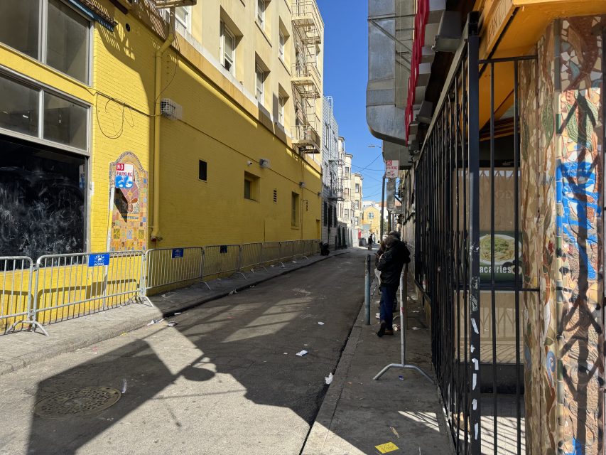 A narrow urban alley with yellow buildings on the left and a person standing by metal barricades. The alley is littered with debris and paper.