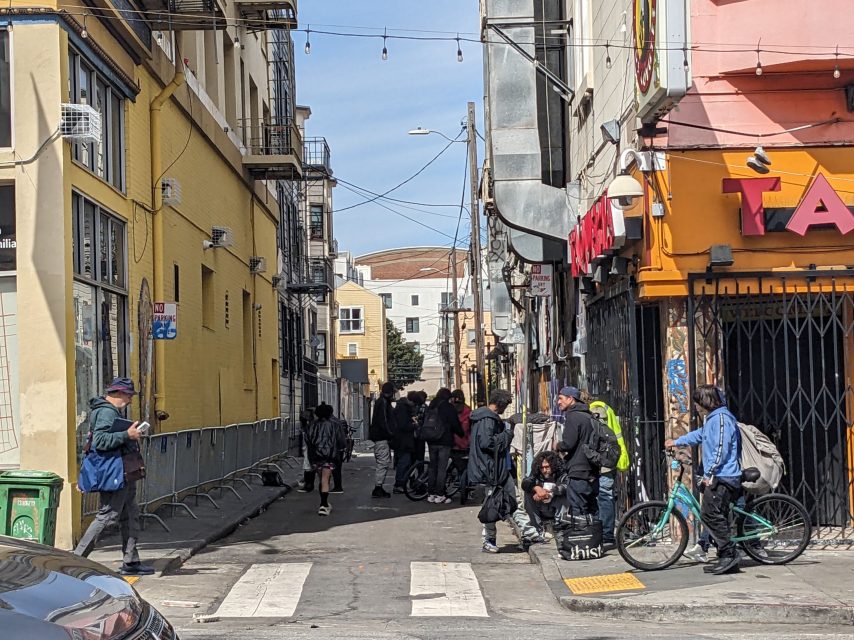 A group of people gathers in a narrow urban alleyway with colorful buildings and string lights. Some individuals stand, others sit on the ground. A person on a bike approaches the intersection.