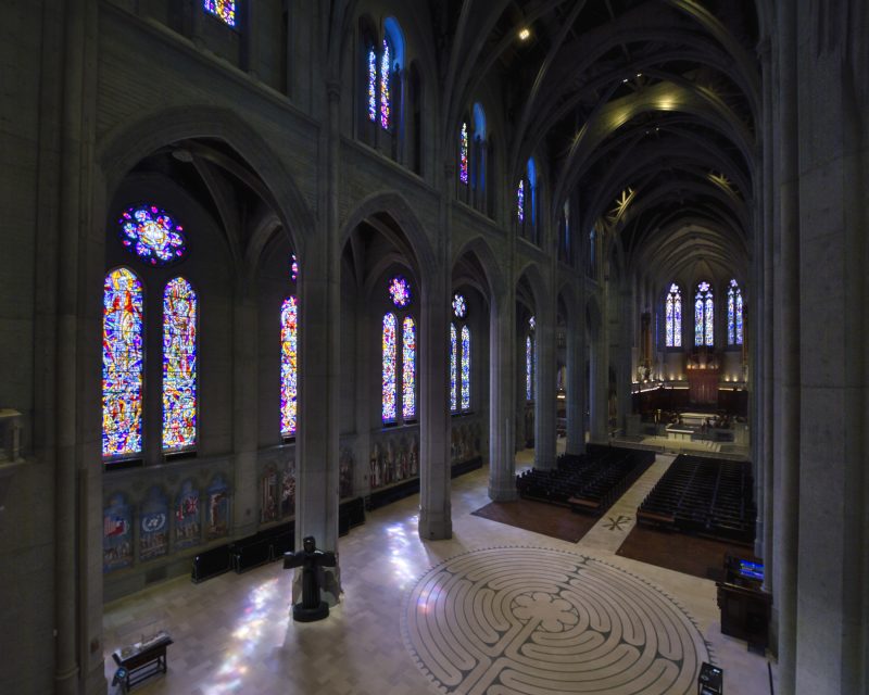 Interior of a large cathedral with tall stained glass windows and stone arches. A labyrinth pattern is on the floor, and rows of pews lead to the altar.