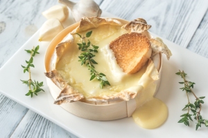 Baked cheese in a round wooden box with a toasted slice on top, surrounded by fresh thyme sprigs on a white plate.