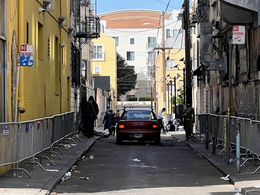 A red car navigates the narrow urban alley of 16th St. Plaza, flanked by yellow and gray buildings. Metal barriers and scattered debris litter the ground.
