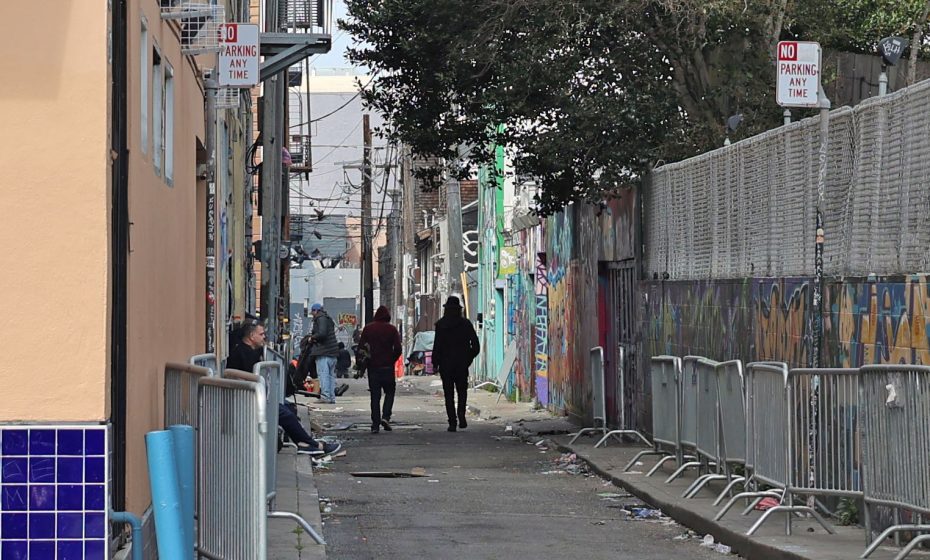 An urban alleyway with graffiti-covered walls. Two people walk down the path, which is lined with metal barriers and scattered trash. Other individuals are seated along the sides.