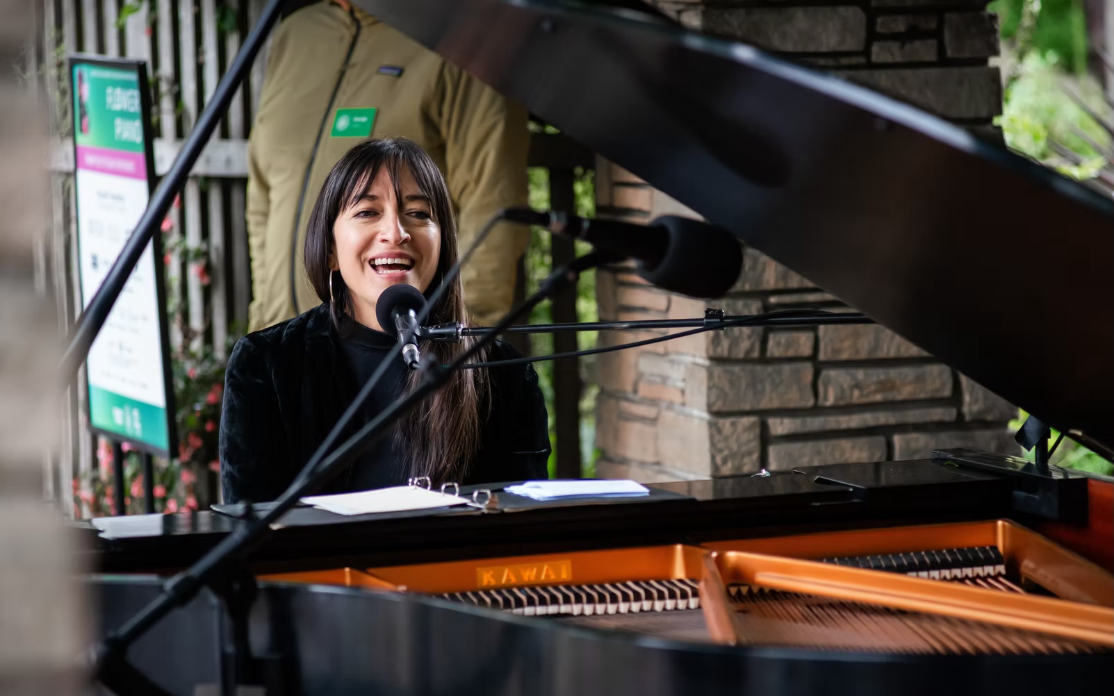 A person singing and playing a grand piano outdoors, with a microphone setup in front of them.
