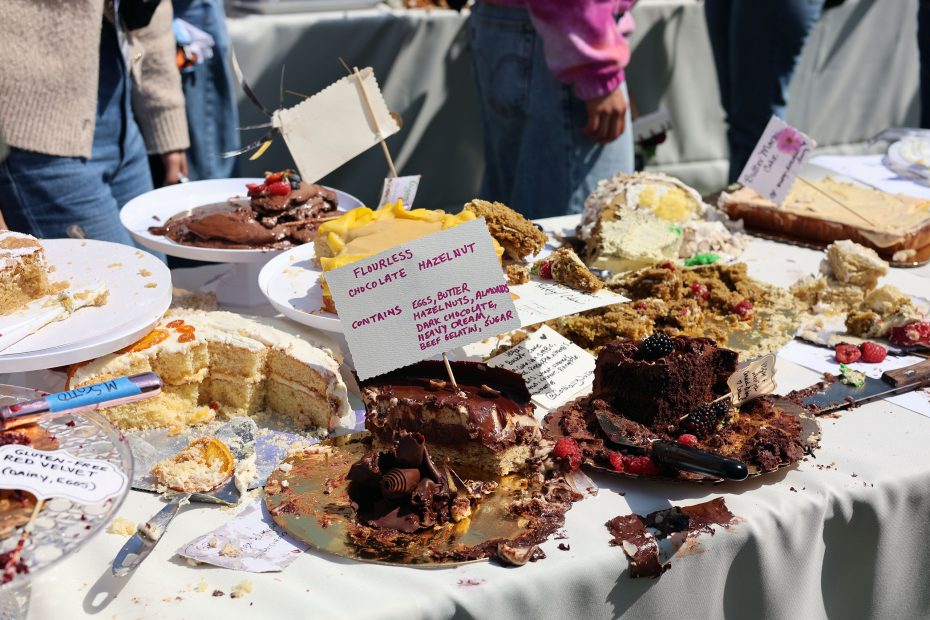 Assorted cakes, some partially eaten, are displayed on a table. Various labels identify the ingredients. A few utensils are scattered among the cakes.