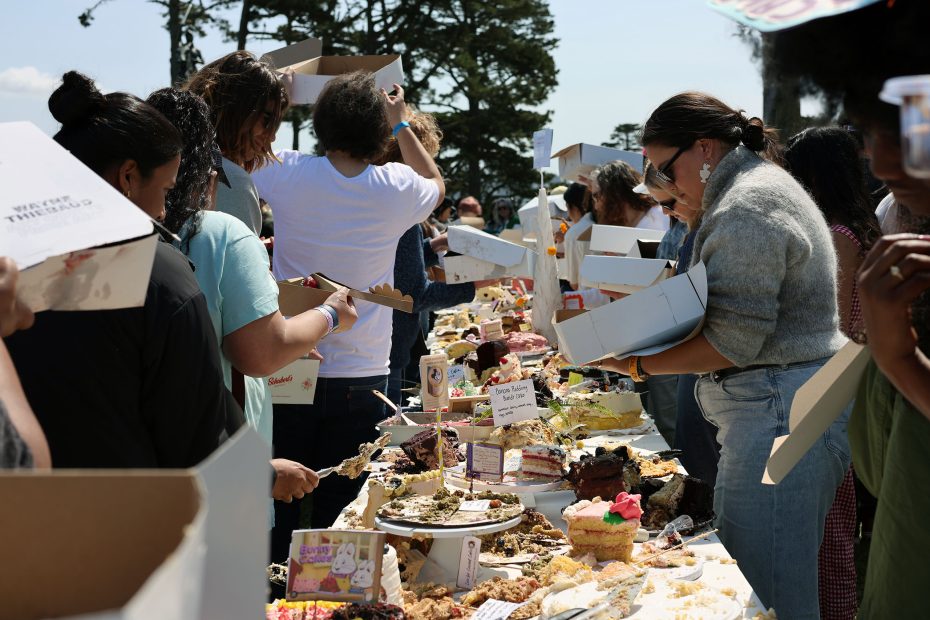 A group of people stands around a long table filled with a variety of cakes and desserts outdoors, each holding boxes or plates, with trees visible in the background.