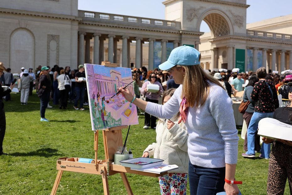 A woman wearing a blue cap paints on an easel outside a large building during a public art event, surrounded by a crowd.