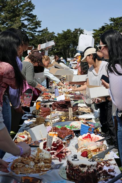 People gather around outdoor tables filled with assorted cakes and desserts, selecting items under a clear sky.