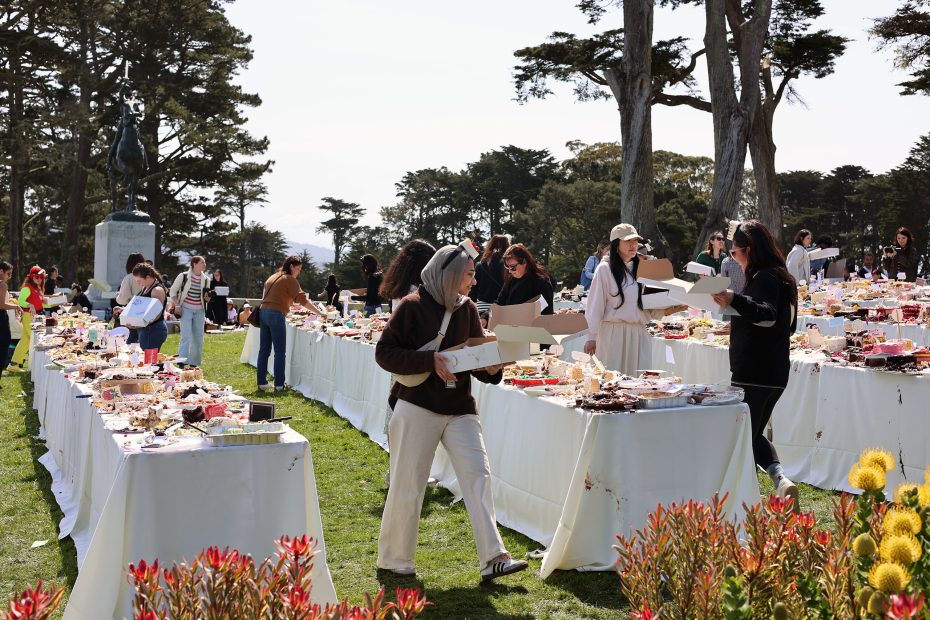 People collecting food from tables covered with various dishes outdoors in a park setting, surrounded by trees and plants.