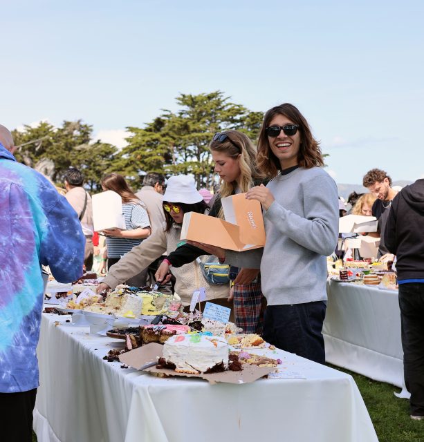 People selecting cakes from tables at an outdoor event. A person holds an orange box and smiles at the camera. Others are browsing the cakes. Trees are visible in the background.