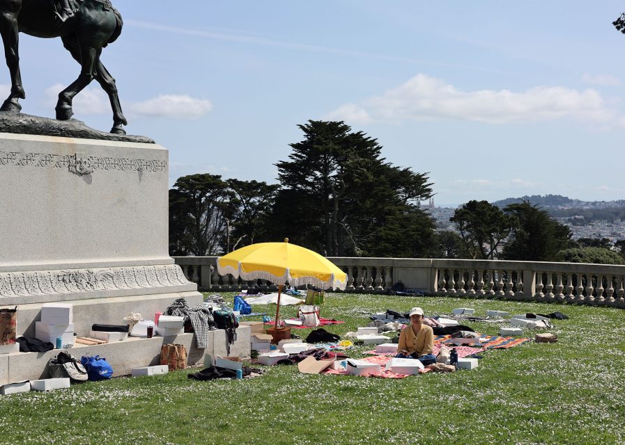 A person sits on a blanket surrounded by boxes and belongings near a statue base in a park. A yellow umbrella provides shade. Trees and a distant cityscape are visible in the background.