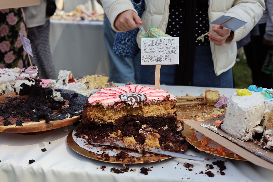 A person is holding a label above partially eaten cakes on display, including a layered cake with chocolate and vanilla sections. Other cake remnants are scattered on the table.