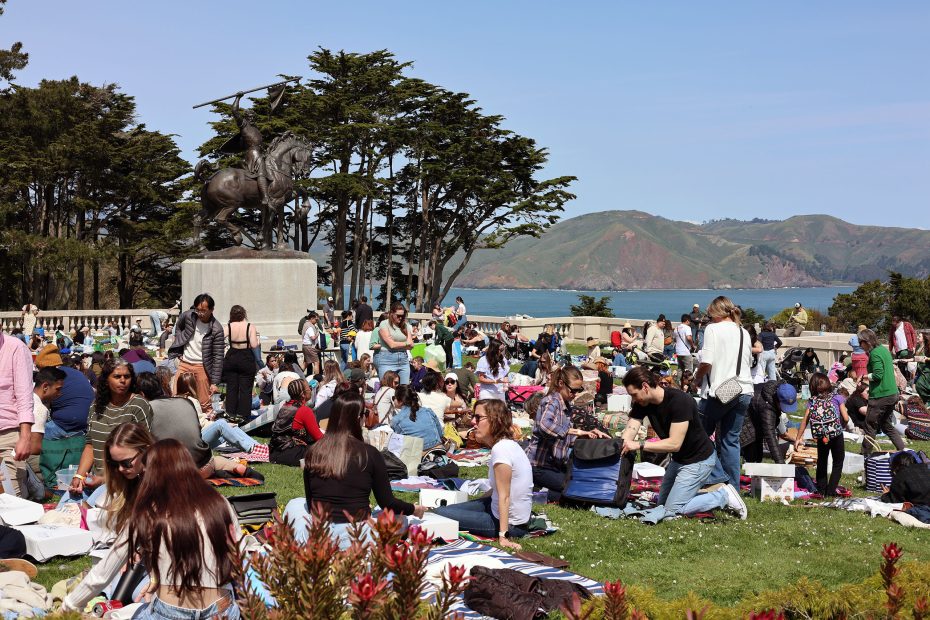 A large group of people gather on a grassy area near a statue, with a scenic view of hills and water in the background.