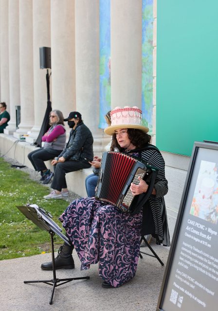 A person in a pink hat plays an accordion outdoors. They sit in front of a wall with columns. Other people are seated in the background.