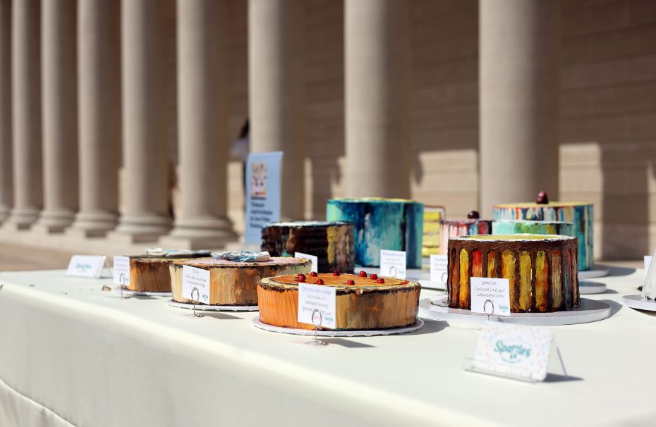 A row of artistically decorated cakes displayed on a table outdoors, each labeled with information. Columns of a building are visible in the background.