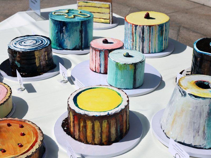 Assorted colorful cakes on display with various geometric designs and patterns, arranged on a white tablecloth outdoors.