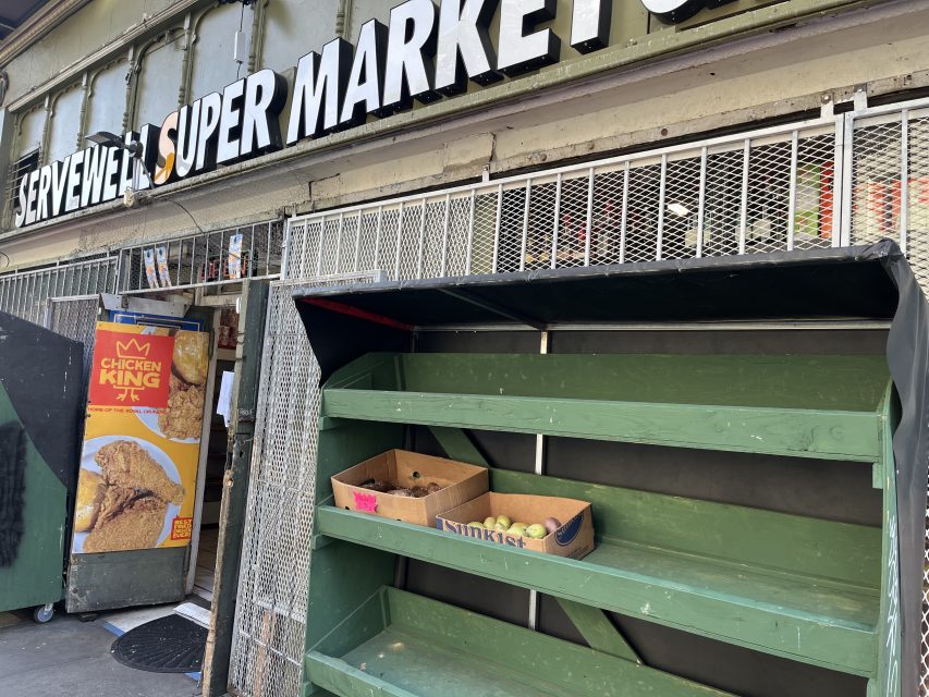 Empty green produce shelves outside a supermarket with a few apples in a box. A poster of fried chicken is visible next to the entrance.