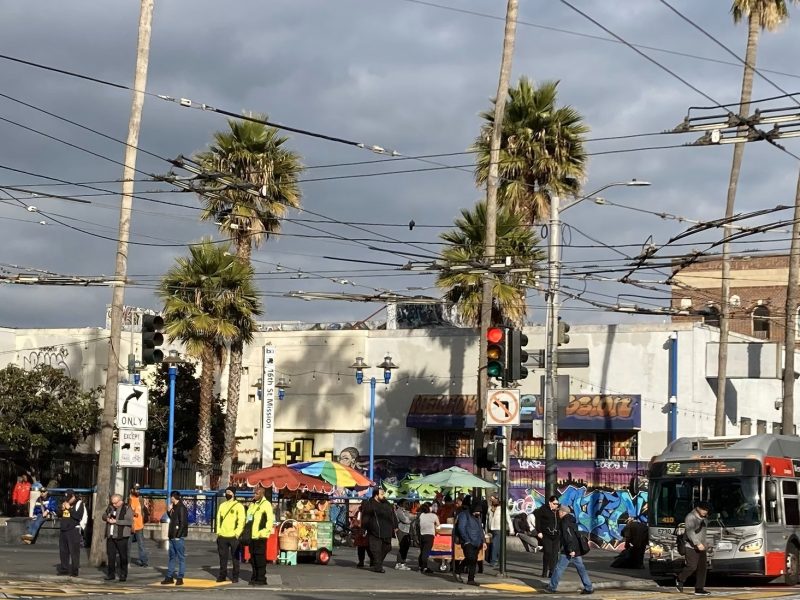 Street scene with palm trees, graffiti-covered buildings, overhead wires, a bus, and people near a street vendor. Overcast sky visible.