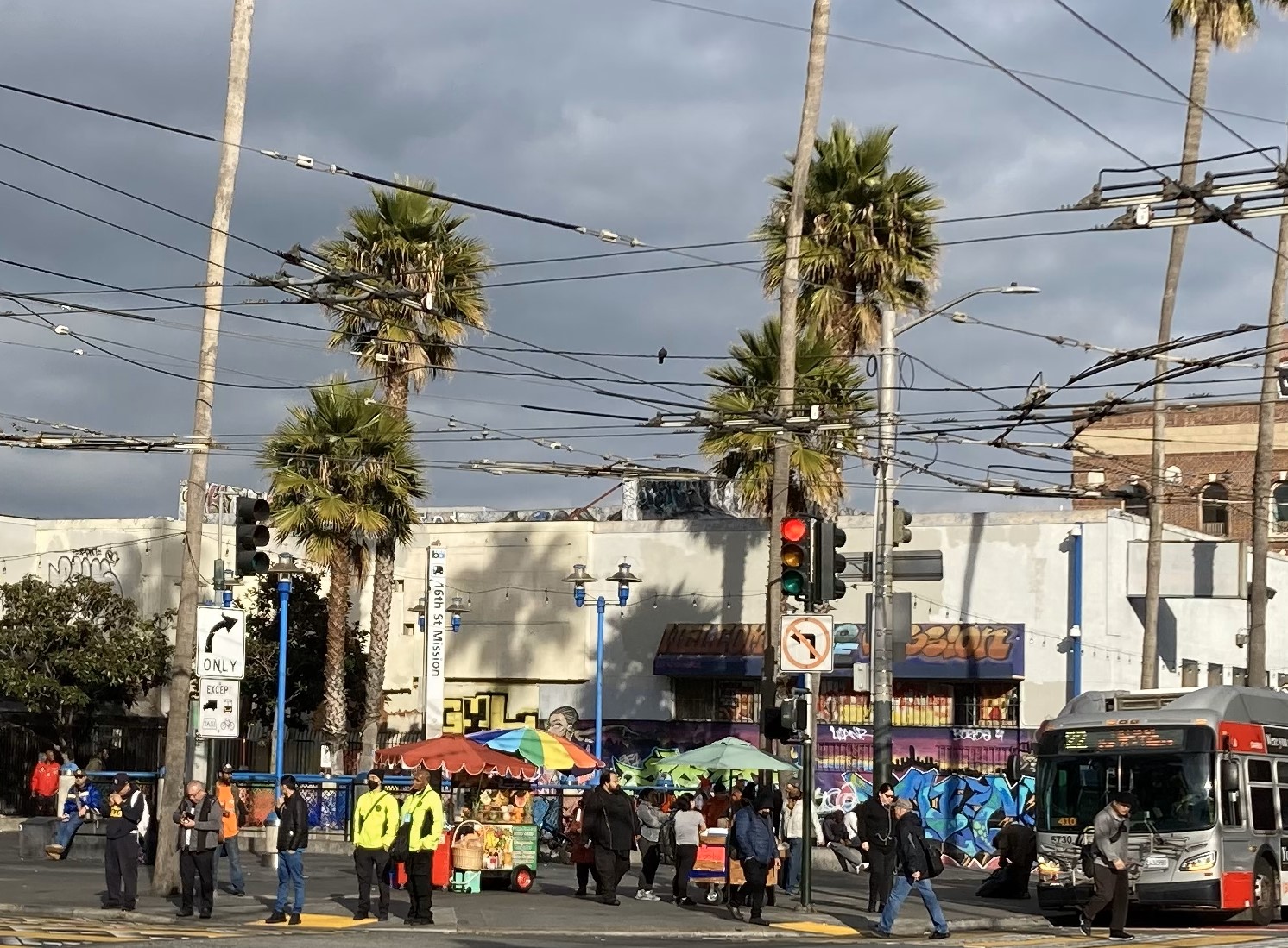 Street scene with palm trees, a bus, people, and a hot dog stand under a cloudy sky. Graffiti on buildings and traffic lights are visible.