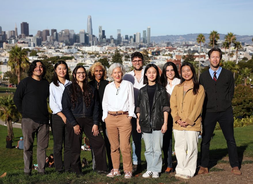 A group of ten people standing together on a grassy area with a city skyline in the background.