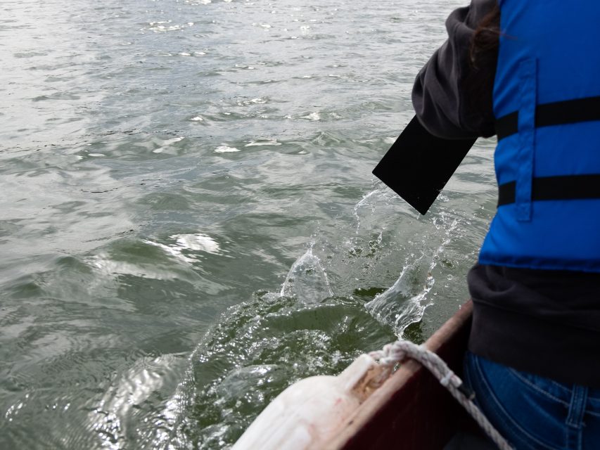Person in a blue life jacket rowing a boat on water with a paddle, causing splashes.