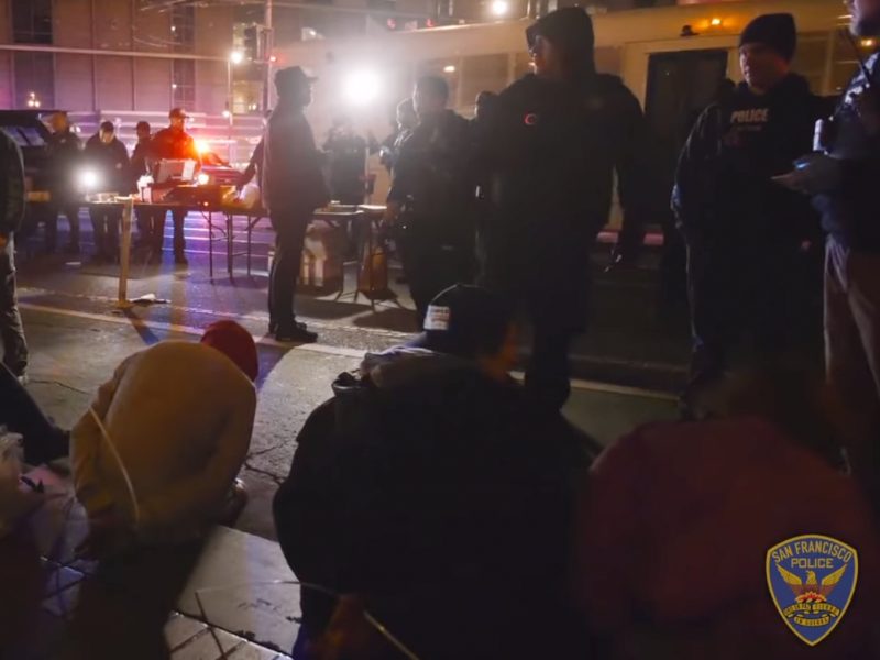 Group of police officers and individuals on a city street at night. Officers are interacting with seated individuals. Police emblem visible in the foreground.