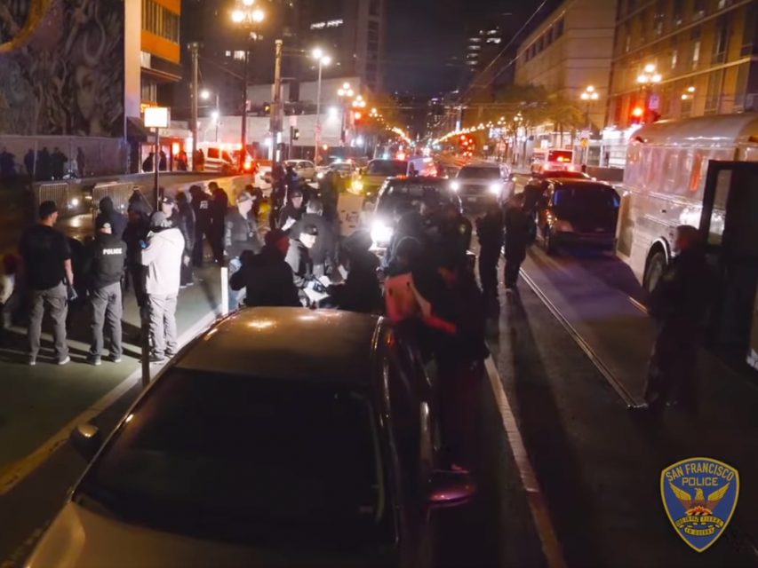 A nighttime street scene with multiple police officers and vehicles. The street is crowded with people, and city buildings are visible in the background. San Francisco Police Department badge displayed.