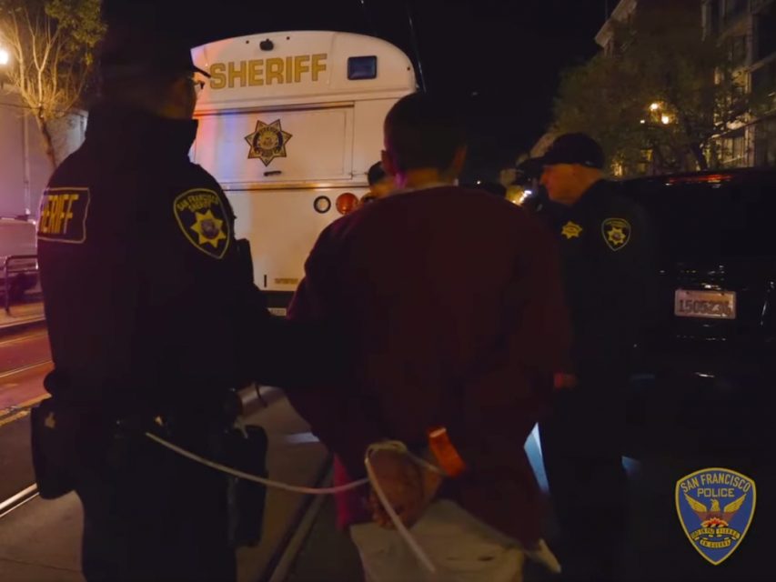 A person with hands zip-tied behind their back stands with two uniformed officers in front of a vehicle marked "Sheriff." The San Francisco Police Department emblem is visible in the corner.